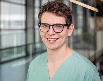 Ein lächelnder Mann mit Brille und grüner medizinischer Kleidung steht in einem modernen Gebäude mit Glaswänden im Hintergrund.