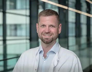 Ein lächelnder Mann in einem weißen Kittel steht in einem modern gestalteten Bürogebäude mit Glaswänden im Hintergrund.