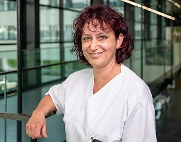 Eine lächelnde Person in einem weißen Kittel steht in einem modernen Glasgebäude. Die Umgebung wirkt professionell und ordentlich. Tageslicht scheint herein.