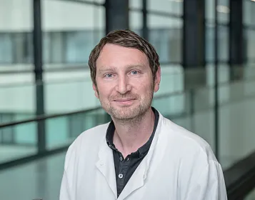 Ein Mann im weißen Kittel steht in einem modernen Gebäude mit verglasten Wänden. Er hat kurzes, braunes Haar und lächelt in die Kamera.