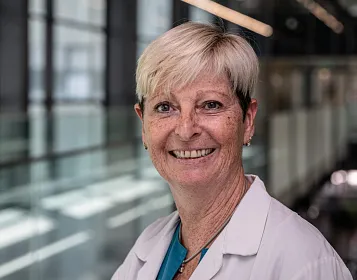 Ältere Frau mit kurzem weißem Haar lächelt in die Kamera. Sie trägt einen weißen Kittel und steht vor einem modernen, lichtdurchfluteten Gebäude.