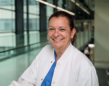 Eine Frau in einem weißen Arztkittel steht in einem modernen, hellen Gebäude. Sie lächelt und trägt darunter blaue Kleidung. Im Hintergrund sind Fenster zu sehen.