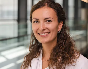 Eine Frau mit lockigem, braunem Haar lächelt in die Kamera. Sie trägt einen weißen Blazer. Im Hintergrund sind Fenster und ein heller Raum zu sehen.