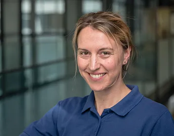 Eine lächelnde Frau in einem blauen Poloshirt steht in einem modernen, lichtdurchfluteten Gebäude mit Glaswänden. Der Hintergrund ist verschwommen.