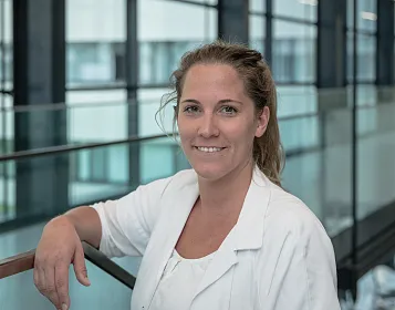 Person in a white lab coat smiles in a modern building with glass walls.