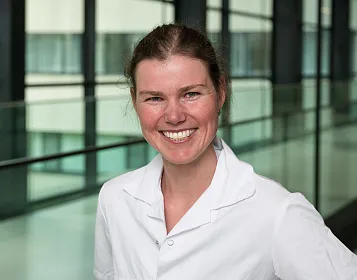 Eine Person in einem weißen Laborkittel lächelt in die Kamera. Der Hintergrund zeigt moderne Glas- und Stahlarchitektur mit großen Fenstern.