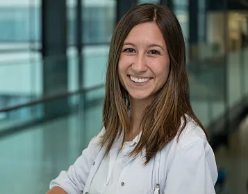 Lächelnde Frau in weißem Kittel steht in einem modernen Gebäude mit Glaswänden. Sie schaut direkt in die Kamera, im Hintergrund sind unscharfe Büroumgebungen.