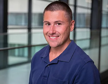 Porträt eines lächelnden Mannes in einem dunkelblauen Hemd in einem modernen Bürogebäude mit Glaswänden im Hintergrund.