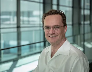 Ein lächelnder Mann mit Brille und hellem Hemd steht vor einer großen Glasfassade. Der Hintergrund ist modern und hell beleuchtet.