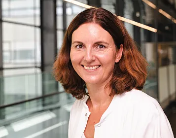 Eine Frau in weißem Kittel lächelt freundlich in die Kamera. Sie befindet sich in einem modernen Gebäude mit Glas- und Metallakzenten und großen Fenstern.