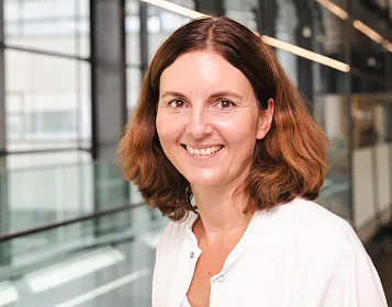 Eine Frau in weißem Kittel lächelt freundlich in die Kamera. Sie befindet sich in einem modernen Gebäude mit Glas- und Metallakzenten und großen Fenstern.