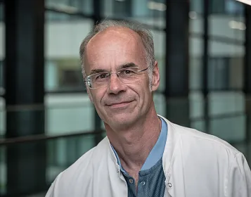 Ein älterer Mann mit Brille und weißen Haaren trägt einen weißen Kittel und steht in einem modernen Gebäude mit Glaswänden und weicher Beleuchtung.