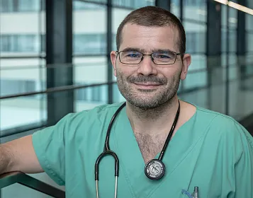 Ein Mann in grüner medizinischer Kleidung steht in einem modernen Flur. Er trägt eine Brille und ein Stethoskop um den Hals. Der Hintergrund ist verglast.