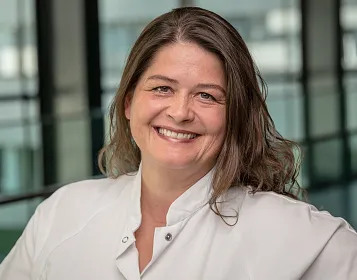 Porträt einer lächelnden Person in einem weißen Kittel vor einem verschwommenen Hintergrund mit großen Glasfenstern. Die Atmosphäre wirkt professionell.