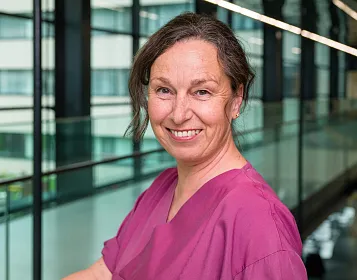 Eine lächelnde Frau in pinker Krankenhauskleidung steht in einem modernen Gebäude mit Glaswänden im Hintergrund. Sie trägt bräunliches Haar zurückgebunden.