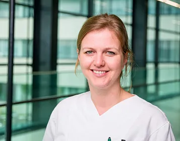 Eine Frau in weißer Arbeitskleidung lächelt in einem modernen Bürogebäude mit viel Glas und Licht. Im Hintergrund sind große Fenster und ein Geländer sichtbar.