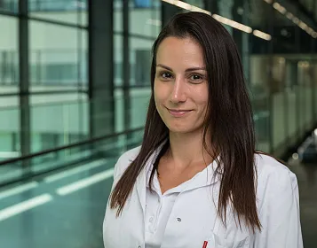 Eine Person in einem weißen Laborkittel steht in einem modernen Gebäude mit großen Glasfenstern. Sie lächelt und hat lange, dunkelbraune Haare.