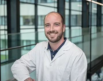 Ein lächelnder Mann im weißen Laborkittel steht in einem modernen Gebäude mit Glaswänden. Die Umgebung wirkt professionell und hell beleuchtet.