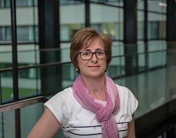 Eine Person mit Kurzhaarschnitt und Brille trägt ein weißes T-Shirt und einen rosa Schal. Sie steht in einem modernen Gebäude mit Glaswänden.