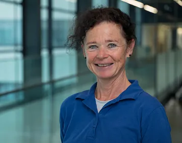 Eine Frau mittleren Alters steht in einem modernen Gebäude mit Glaswänden. Sie trägt ein blaues Hemd und lächelt freundlich in die Kamera.