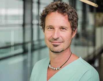 Ein lächelnder Mann in medizinischer Kleidung steht in einem modernen Gebäude. Der Hintergrund zeigt Glaswände mit unscharfen Strukturen.