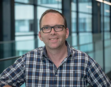 Ein lächelnder Mann trägt ein kariertes Hemd und eine Brille. Er steht in einem modernen Gebäude mit Glaswänden und hellblauer Beleuchtung im Hintergrund.