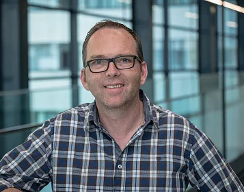 Ein Mann mit Brille und kariertem Hemd steht in einem modernen Gebäude mit großen Fenstern. Das Ambiente ist hell und minimalistisch.