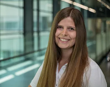 Porträt einer lächelnden Person mit langen, glatten Haaren in einem modernen Bürogebäude mit Glaswänden und heller Beleuchtung im Hintergrund.
