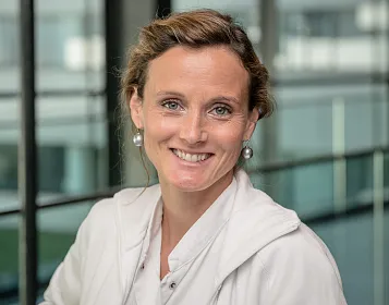 Porträt einer lächelnden Person in einem modernen Büro mit großen Fenstern im Hintergrund. Die Person trägt helle Kleidung und auffällige Ohrringe.