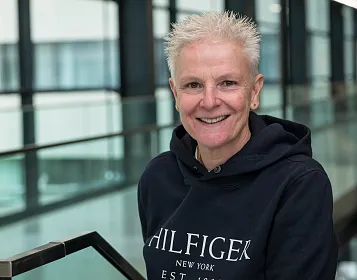 Eine Person mit kurzen weißen Haaren und dunklem Hoodie steht in einem modernen Gebäudeflur mit Glasgeländern und lächelt in die Kamera.