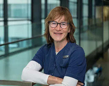 Porträt einer lächelnden Person in medizinischer Kleidung und Brille, die in einem modernen Gebäude mit Glaswänden steht, mit verschränkten Armen.