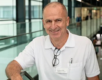 Ein älterer Mann mit kurzem Haar und Brille lächelt in die Kamera. Er trägt ein weißes Hemd und steht in einem modernen Gebäude mit Glaswänden.