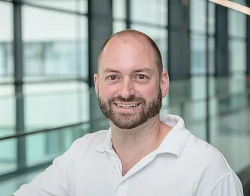 Lächelnder Mann mit Glatze und Vollbart in einem weißen Hemd, steht in einem modernen Büro mit Glaswänden und großen Fenstern im Hintergrund.