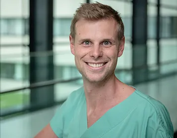 Ein lächelnder Mann in grüner Krankenhauskleidung steht vor einem Fenster in einem modernen Gebäude. Im Hintergrund sind verschwommene Glasstrukturen zu sehen.