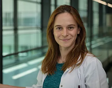 Eine lächelnde Person in einem weißen Kittel steht in einem modernen Gebäude mit Glaswänden. Sie hat lange, braune Haare und trägt ein blaues Oberteil.