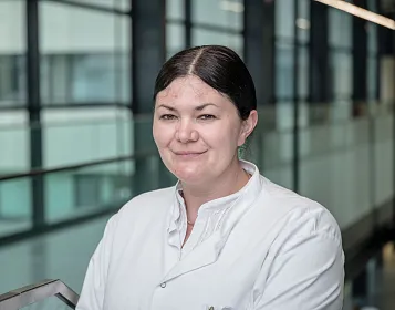 Eine Person in einem weißen Laborkittel steht in einem modernen Gebäude mit Glaswänden. Sie hat dunkle, nach hinten gebundene Haare und lächelt leicht.