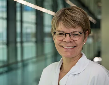 Eine lächelnde Person mit Kurzhaarfrisur und Brille, in einem weißen Laborkittel, vor einem großen Fenster mit moderner Architektur im Hintergrund.