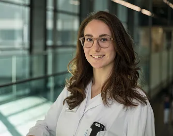 Frau im weißen Laborkittel steht vor einem Glasgeländer in einem modern ausgestatteten Gebäude mit natürlichem Licht. Sie trägt eine Brille und lächelt leicht.