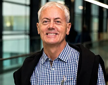 Ein Mann mittleren Alters mit grauen Haaren und kariertem Hemd lächelt in einem modernen, gläsernen Gebäude, das einen hellen und offenen Eindruck vermittelt.