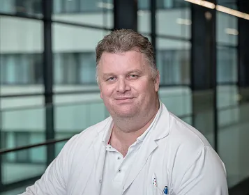 Porträt eines Mannes mittleren Alters, der in einem weißen Arztkittel lächelt. Hinter ihm sind moderne Bürogebäude mit großen Glasfenstern sichtbar.