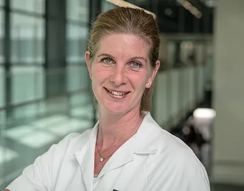 Frau in weißem Arztkittel steht in einem modernen Gebäude mit Glaswänden und lächelt in die Kamera. Im Hintergrund sind verschwommene Korridore sichtbar.