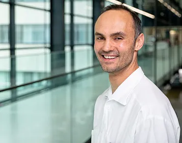 Ein Mann in einem weißen Hemd lächelt in die Kamera. Im Hintergrund sind verschwommene Glaswände und Fenster eines modernen Gebäudes sichtbar.