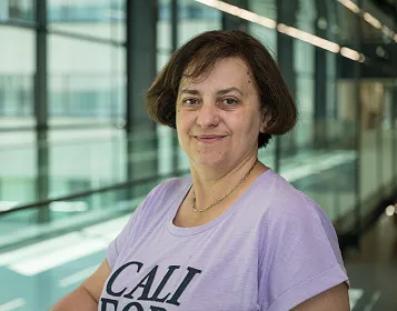 Eine Person mit schulterlangem braunem Haar trägt ein lila T-Shirt mit Schriftzug. Der Hintergrund ist ein modernes Gebäude mit Glaswänden.