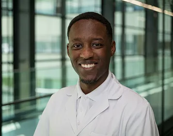 Ein lächelnder Mann in einem weißen Laborkittel steht in einem modernen Gebäude mit Glaswänden und natürlichem Licht im Hintergrund.