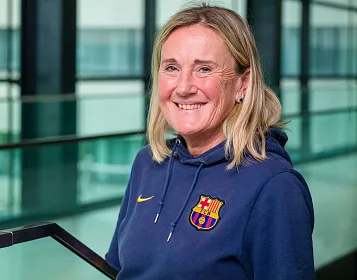 Eine Person mit blonden Haaren lächelt in die Kamera. Sie trägt einen blauen Hoodie mit einem Emblem. Im Hintergrund sind Glaswände zu sehen.