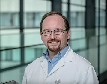 Ein Mann mit Brille und Bart trägt einen weißen Kittel und lächelt in die Kamera. Er steht in einem modernen Gebäude mit großen Fenstern im Hintergrund.