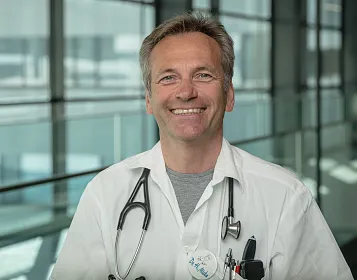 Ein lächelnder Mann in einem weißen Arztkittel mit Stethoskop im modernen Gebäude. Tageslicht fällt durch große Fenster im Hintergrund.