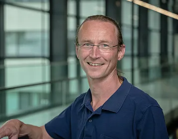 Ein lächelnder Mann mit Brille und langen Haaren steht in einem modernen Gebäude mit großen Glasfenstern. Er trägt ein blaues Polohemd.