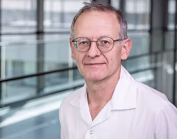 Ein Mann in einem weißen Kittel steht in einem modernen Gebäude mit Glaswänden und natürlichem Licht. Er trägt eine Brille und schaut direkt in die Kamera.