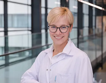Eine freundliche Person mit blonden Haaren und Brille trägt einen weißen Laborkittel. Sie steht in einem modernen Gebäude mit Glaswänden.